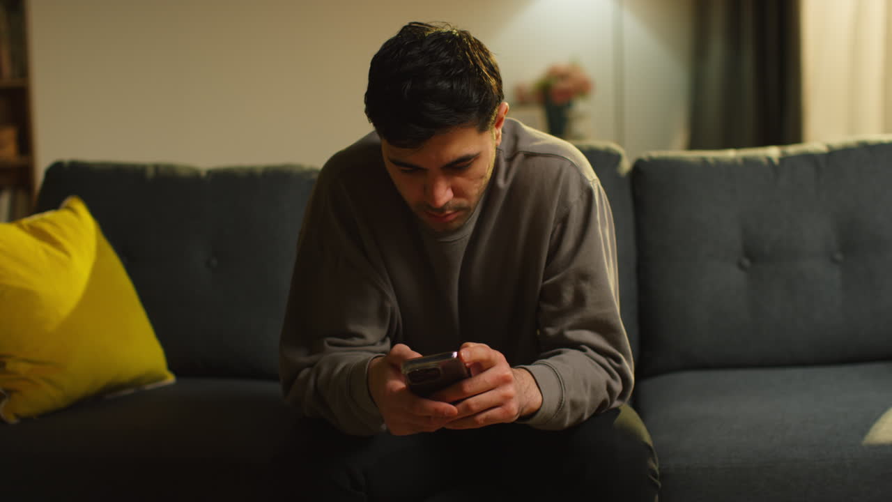 Young Man Spending Evening At Home Sitting On Sofa With Mobile Phone Scrolling Through Internet Or Social Media Streaming Content 3