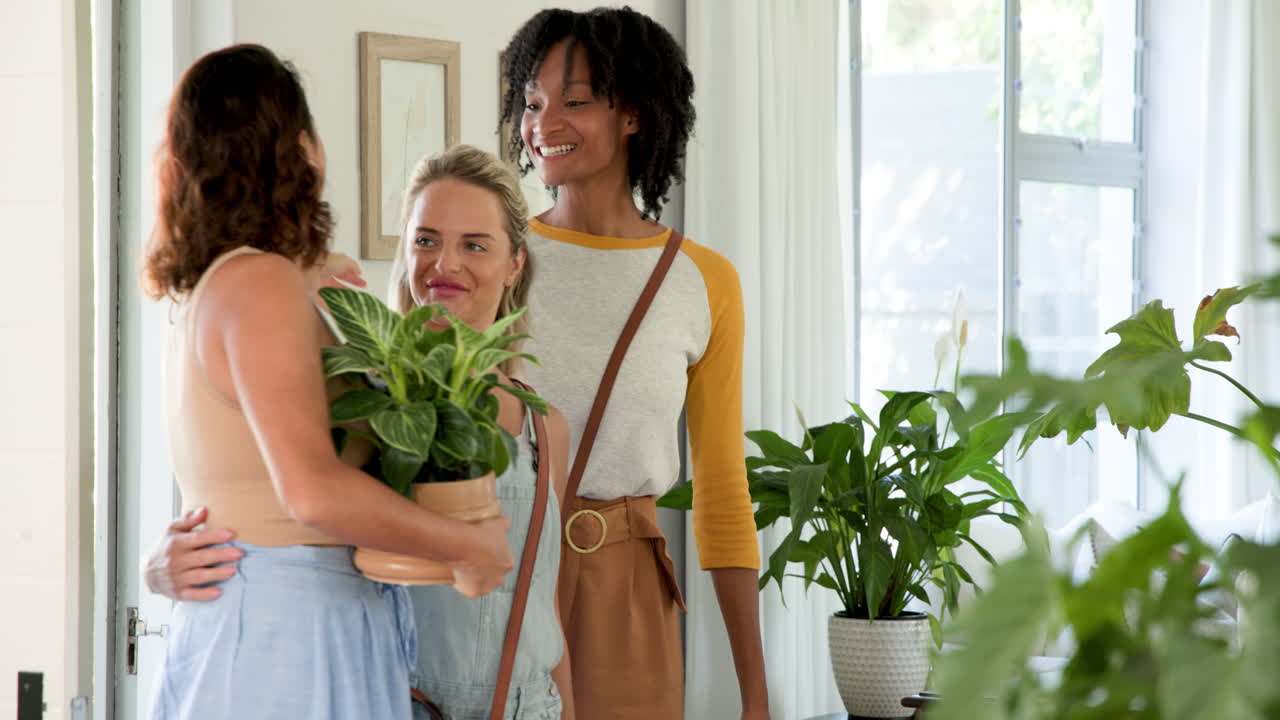 Diverse female friends carrying plants indoors, smiling and enjoying time together at home