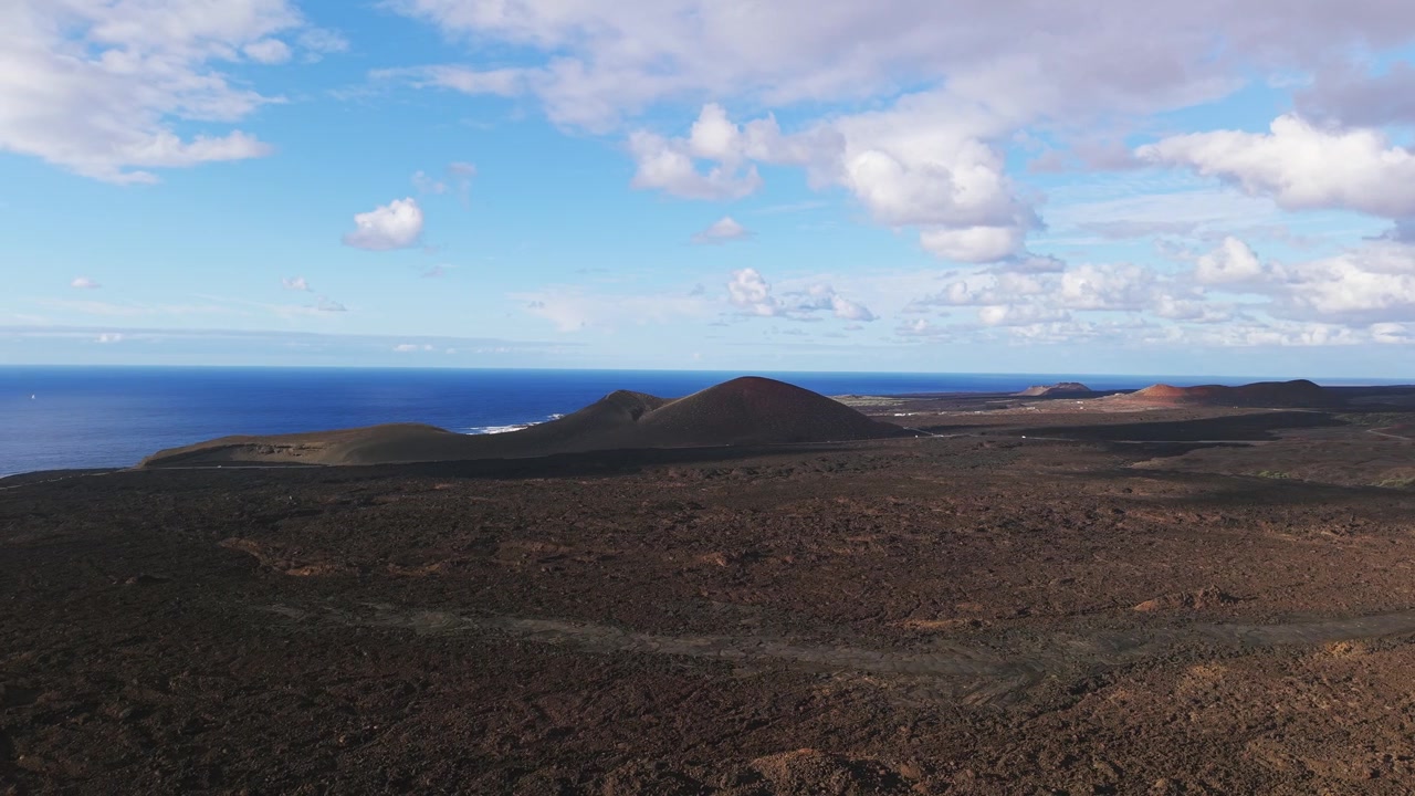 Panorama aereo dei campi vulcanici scuri di Lanzarote, crateri costieri e orizzonte atlantico sotto nuvole sparse