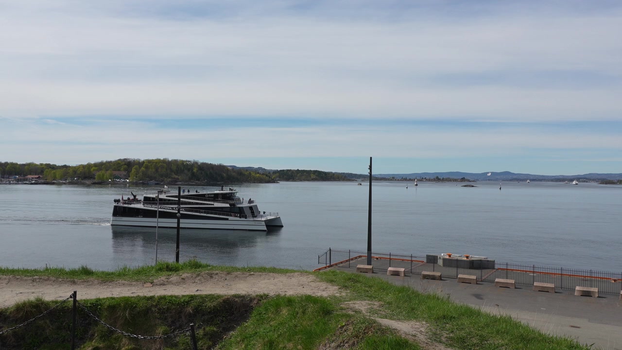 Handhållen film av en elegant båt som glider över Oslos fjordhamn i Norge, inramad av grönt gräs, avlägsna öar och lugnt vatten i mjukt dagsljus med mjuka kamerarörelser.