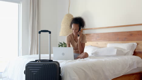 woman on bed using laptop and phone, suitcase nearby, smiling, at home