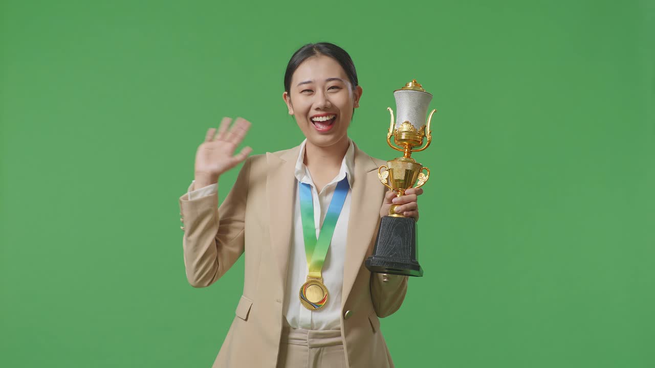 골드 메달과 트로피를 가진 슈트를 입은 아시아 사업가 여성이 스튜디오의 녹색 스크린 배경에서 첫 번째 우승자로 카메라에 손을 흔들고 미소 짓고 있습니다.