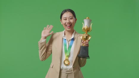 asian business woman in a suit with a gold medal and trophy waving her hand and smiling to camera as the first winner on green screen background in the studio