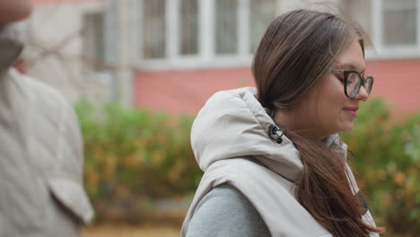 close up partial view of man and lover walking side by side outdoors as woman with glasses and long hair looks at him warmly during stroll with soft smile, background showing trees and building blur