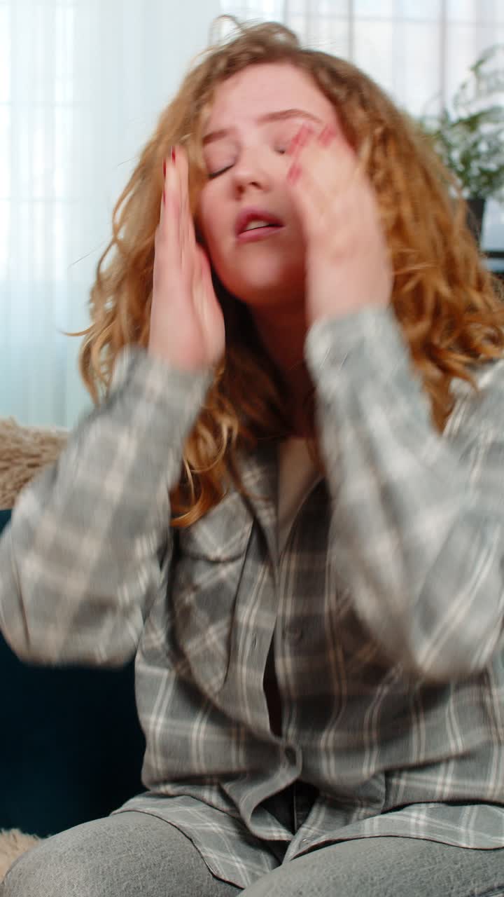 Woman sitting on sofa holding head suffering from strong headache or migraine feeling unwell pain