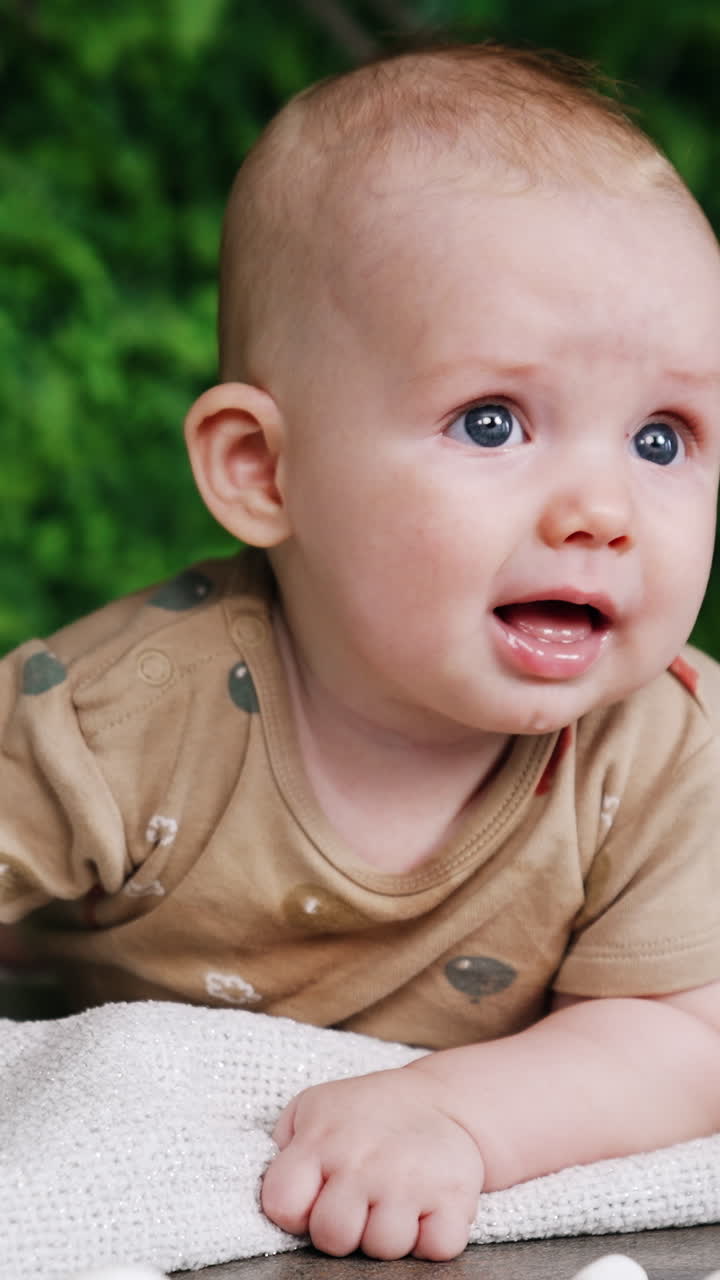 Cute grey-eyed Caucasian baby lying on the belly. Adorable child looking up with interest. Green blurred backdrop. Vertical video