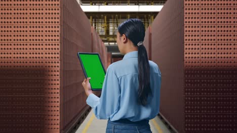 back view of asian female with red brick packed in stacks are stored, checking on tablet with green screen and look at factory