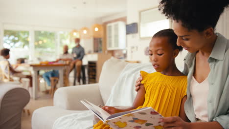 mother and daughter reading book on sofa at home with multi-generation family behind
