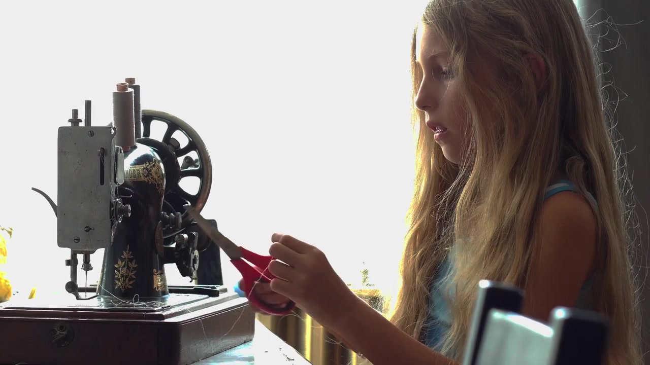 A medium profile static camera shot shows a teen girl operating a vintage hand-crank mechanical sewing machine during a homeschooling craft and practical life skills lesson in Budapest Hungary.