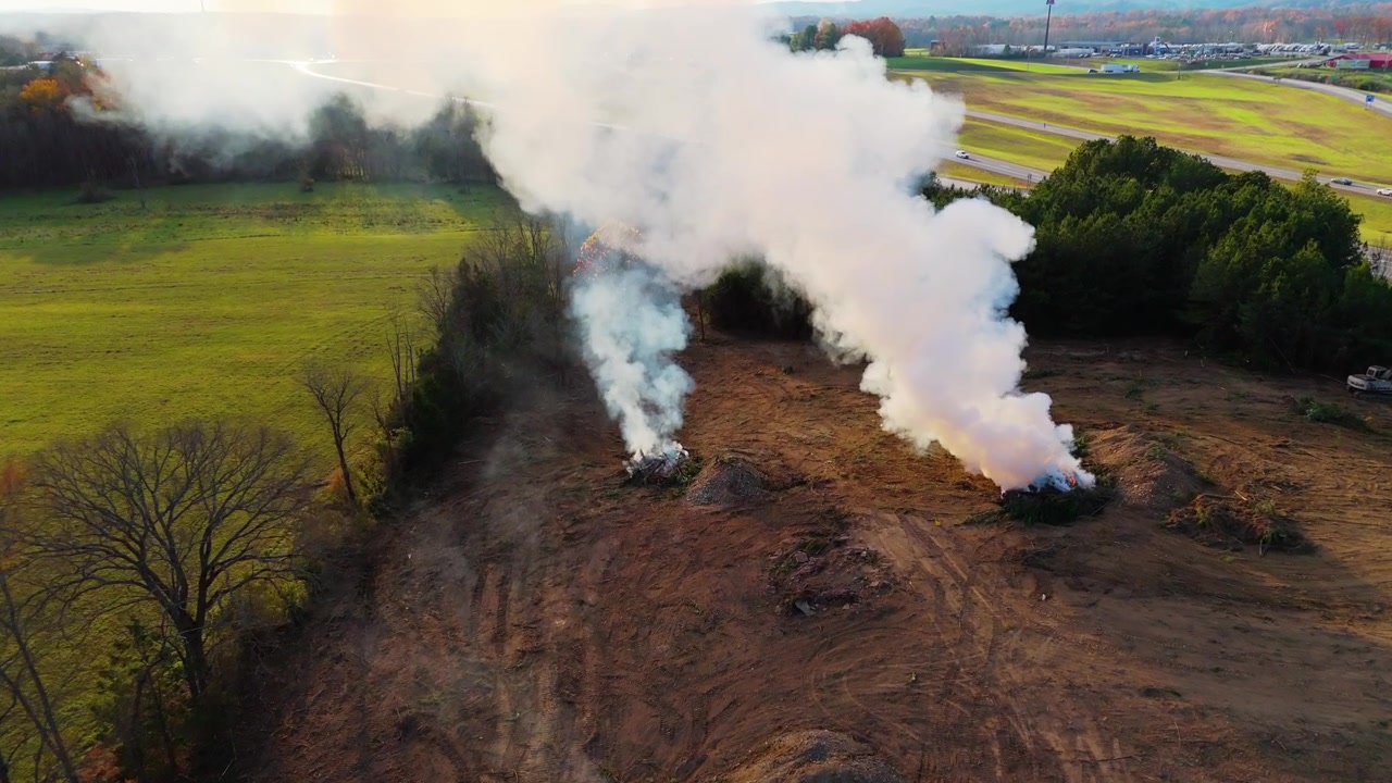 Kırsal Tennessee'de kontrollü bir yakmayı gösteren 4K ağır çekim drone çekimi. Drone dumanın üzerinde uçar ve manzaranın ortasındaki için için yanan çalı yığınlarını ortaya çıkarmak için aşağı doğru döner.