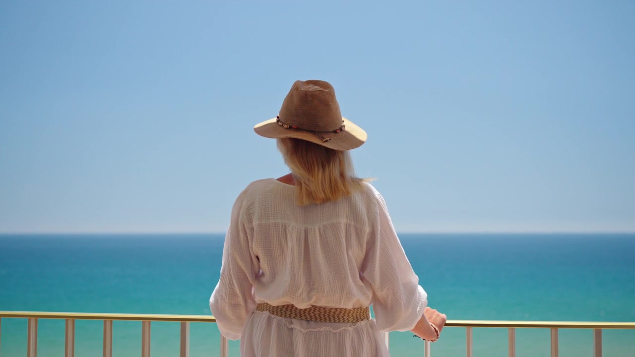 Woman walking onto balcony and looking at calm turquoise sea during sunny summer vacation at beach resort apartment.