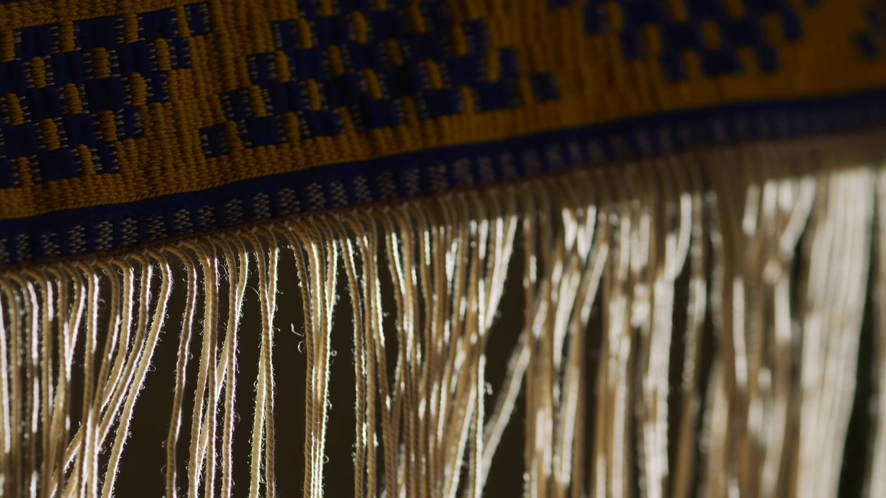 Side lit close view of decorative woven band in mustard yellow and deep blue with long off-white fringe threads, showing rich texture, pattern contrast, and tactile handmade craft, with fringe
