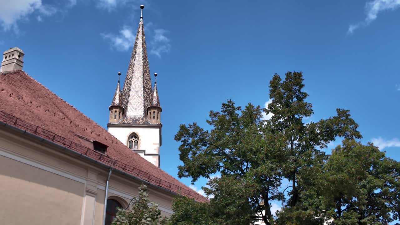 Partial view of the Lutheran Cathedral of Saint Mary in Sibiu, seen behind a historic building and leafy tree in the old town