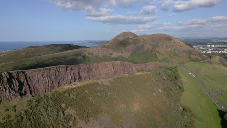 aerial track right to left of arthur's seat grass-covered cliffside and salisbury crags on a blue sky day, edinburgh, scotland, uk