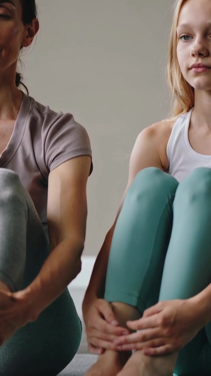 Vídeo vertical: Mulheres adultas e adolescentes de leggings, segurando os joelhos em colchonetes verdes em estúdio, em um momento de calma.