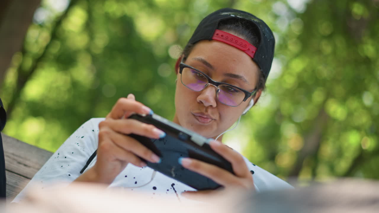 primer plano de una sesión de juego portátil al aire libre: una persona joven con gorra y gafas juega intensamente con una consola en un banco del parque, con un fondo verde borroso, expresión concentrada, luz natural y ropa informal