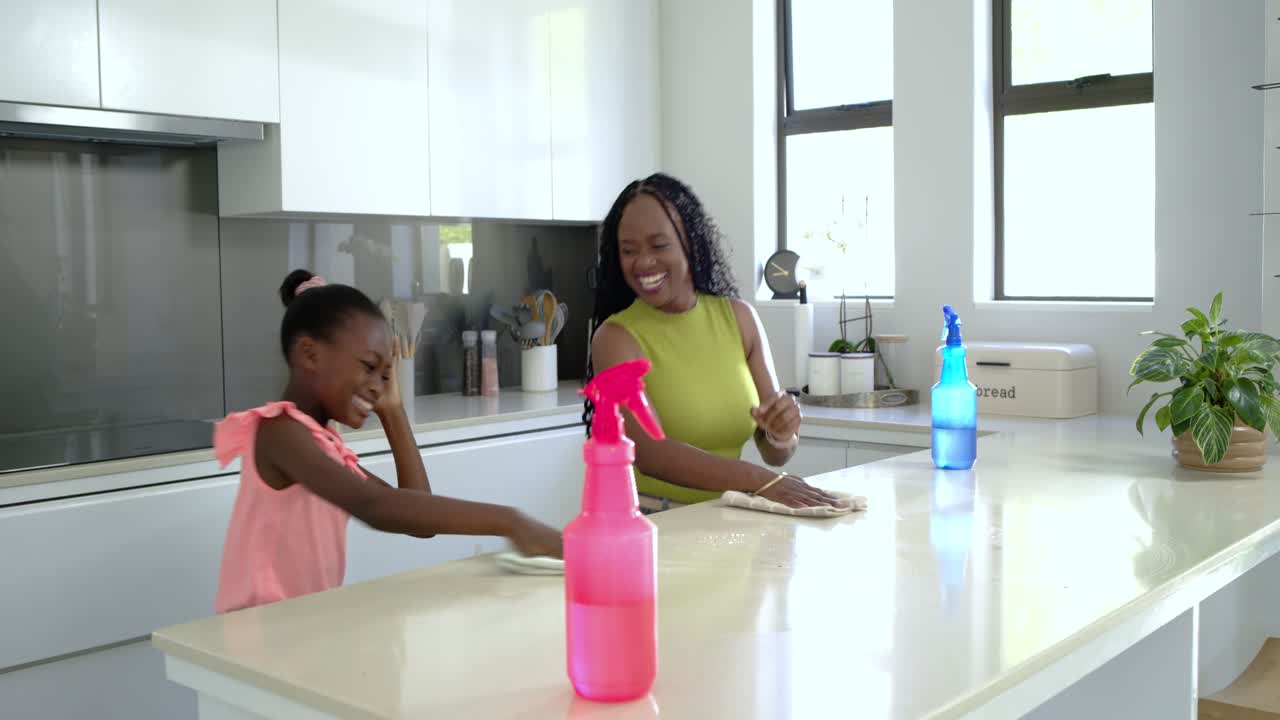 Madre e figlia africane spruzzano e puliscono il piano di lavoro in cucina per rimuovere le briciole con una bottiglia rosa.