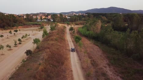 Aerial-done-shot-flying-forward-of-a-grey-car-driving-on-a-gravel-road-with-a-town-at-the-background