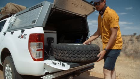 tracking medium hand held shot of a caucasian male tourist in africa as he removes a spare tire from the back of a off-road vehicle and proceeds to roll it to the wheel with a puncture