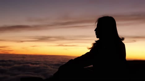 a woman sits in stillness at pico do arieiro, silhouetted against a vivid sunset sky above a sea of clouds.