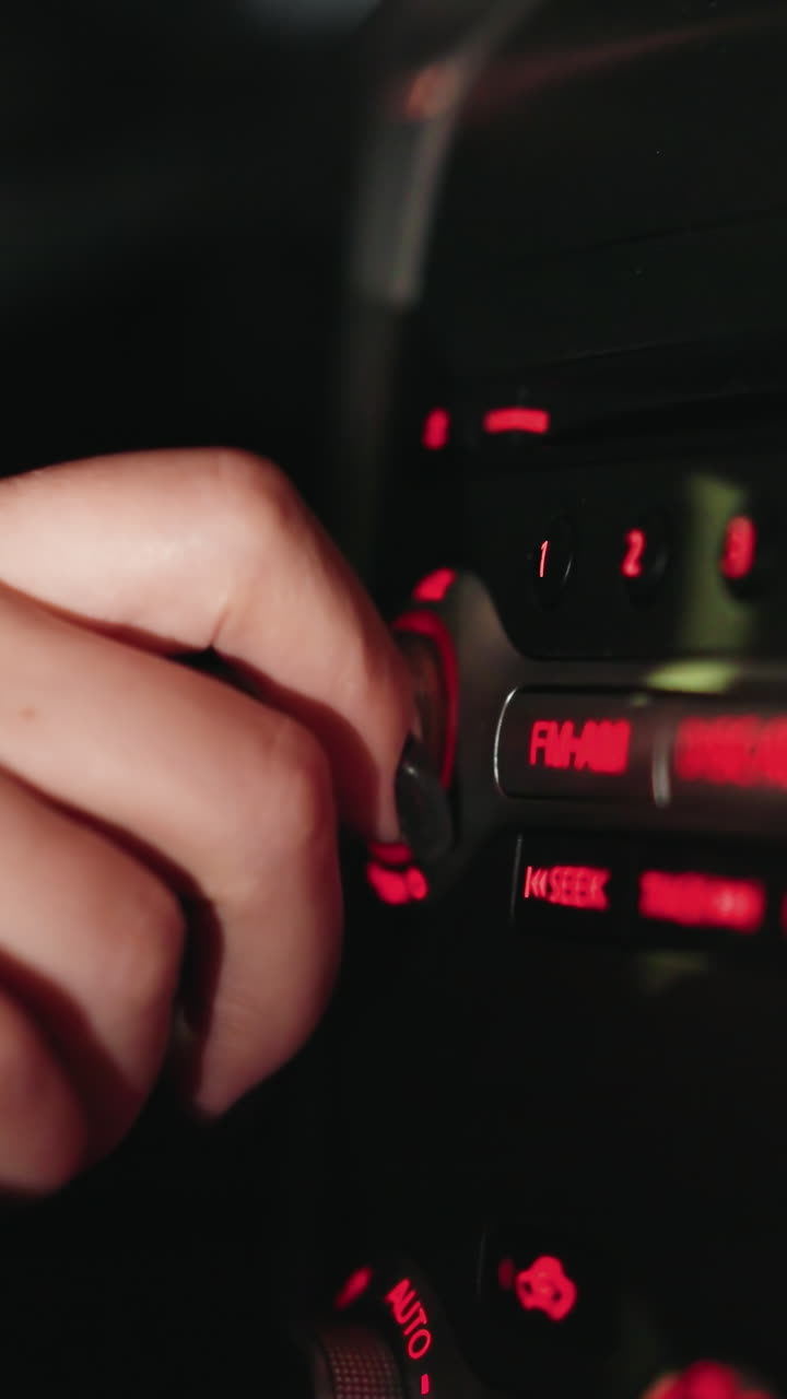 First-person view of a woman's hand turning the FM radio knob inside a car at night. The dashboard is illuminated with red lights