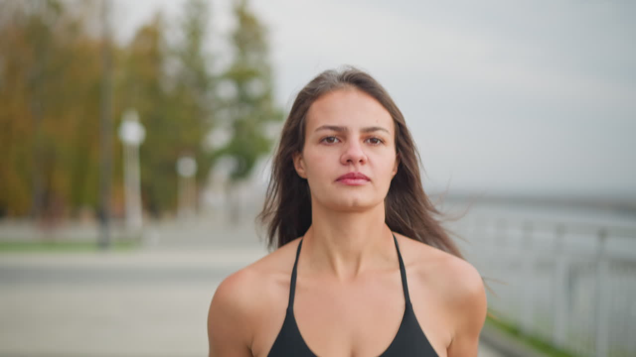 Close-up view of focused young woman in black neck top jogging with full concentration, blurred background showcasing a peaceful park setting, trees, and iron railings