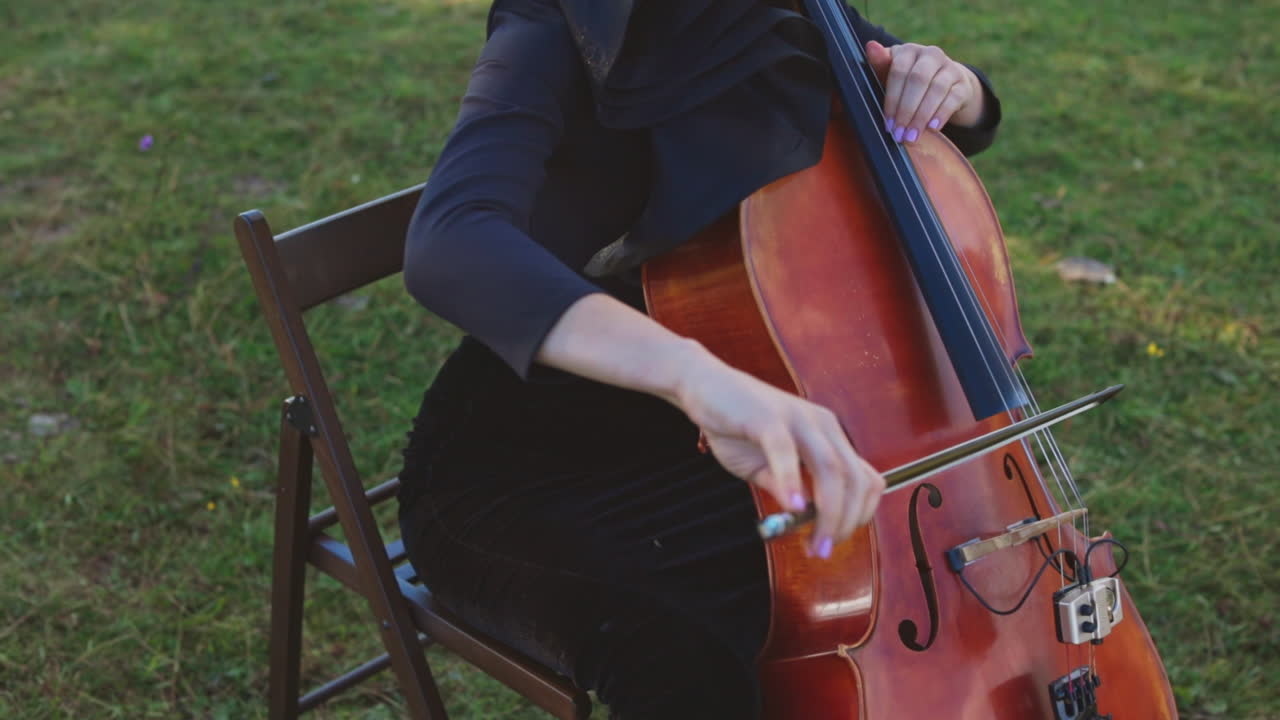 Young woman sitting on the chair at grass lawn playing violoncello. Dark-haired woman performing classical music outdoors.