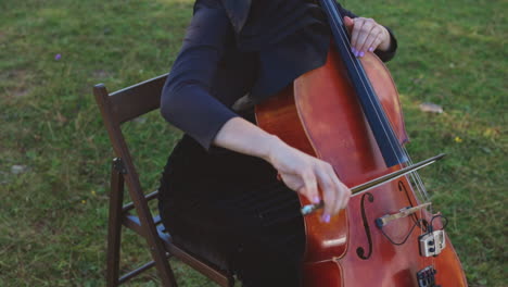 young woman sitting on the chair at grass lawn playing violoncello. dark-haired woman performing classical music outdoors.