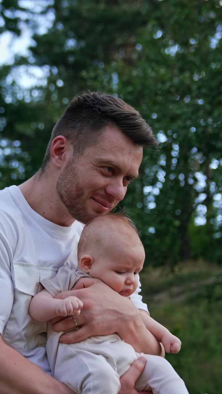 Man waving his baby boy in hands. Caring happy dad kisses child's head. Blurred nature at backdrop. Vertical video