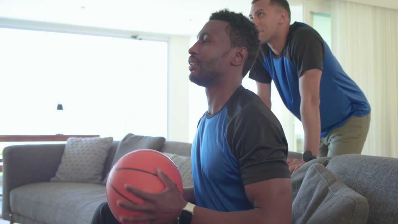 Forskellige mandlige venner kaster basketball, mens de ser en kamp på skærmen og reagerer på et afgørende spil fra sofaen.