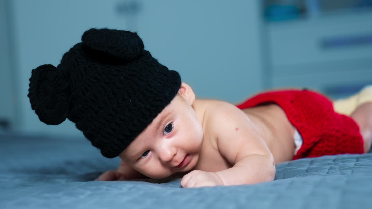 Cute boy in black hat of Mickey Mouse and red pants put on the bed. Small kid strives to rise his head but fails. Close up.