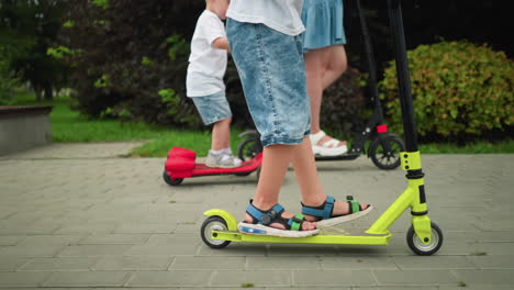 leg view of a woman and her children's legs as they ride scooters, each using their legs to propel forward, the younger child works hard to catch up with the rest