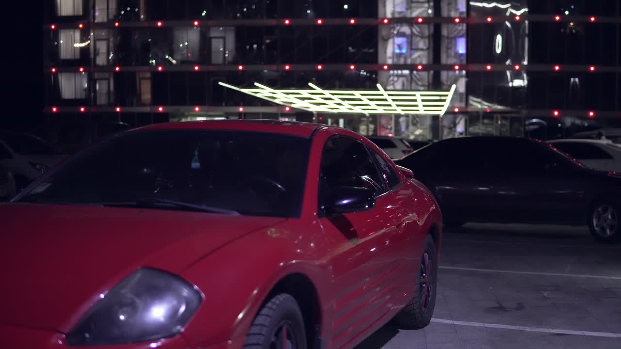 joven sale de un coche rojo por la noche en el estacionamiento y cierra la puerta. vista frontal. activar la señalización