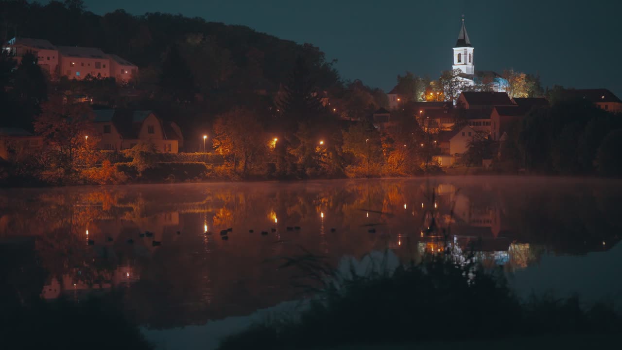 Nighttime footage captures the illuminated St. Sebastian's and Fabian's Church in Prague, its bright lights mirrored on the water