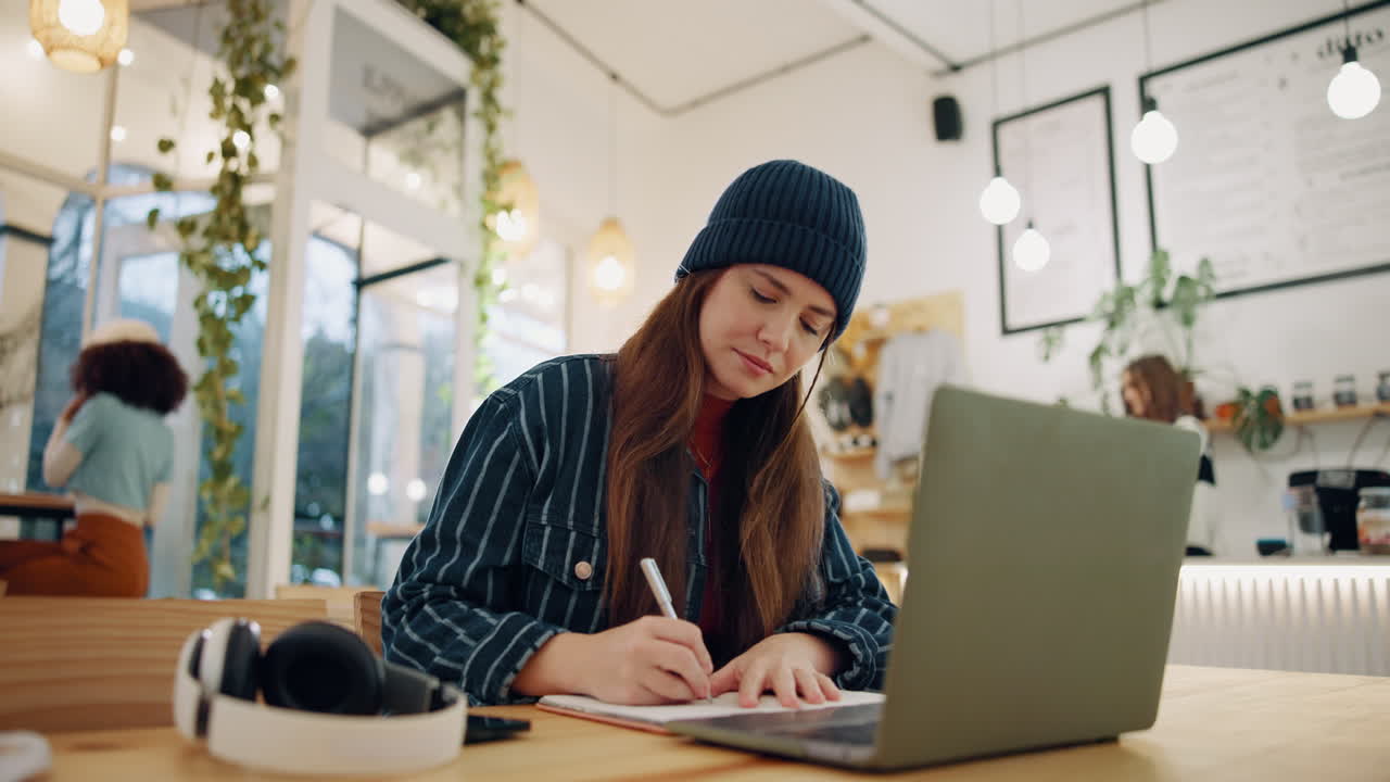Woman writing in a notebook at a cafe with a laptop