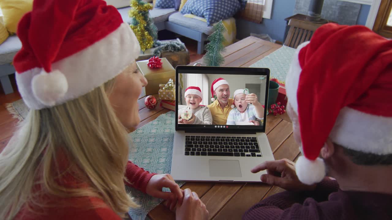 pareja caucásica sonriente con sombreros de santa usando una computadora portátil para una videollamada de navidad con la familia en la pantalla