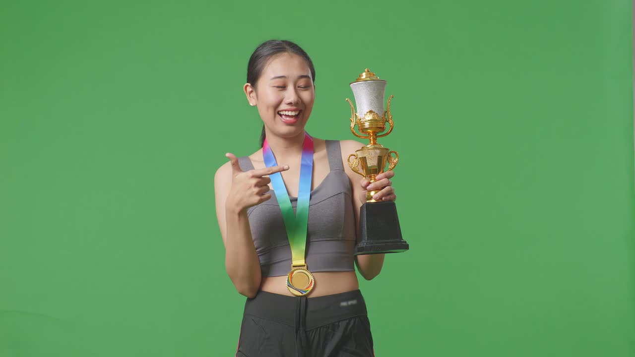 una mujer asiática con una medalla de oro señalando un trofeo de oro en su mano estando orgullosa de ganar como la primera ganadora en el fondo de la pantalla verde en el estudio
