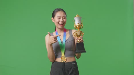 asian woman with a gold medal pointing to a gold trophy in her hand being proud winning as the first winner on green screen background in the studio