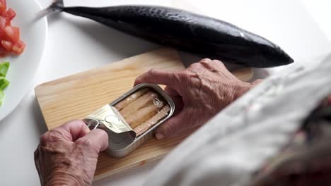 Persona-Caucásica-Con-Manos-Viejas,-Abriendo-Una-Lata-De-Melva,-En-Una-Tabla-De-Madera,-Pescado,-Pimiento-Y-Tomate-En-El-Fondo