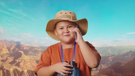 asian boy with a hat and binoculars talking on smartphone while traveling at the top of mountain. boy researcher examines something, travel tourism adventure concept, close up