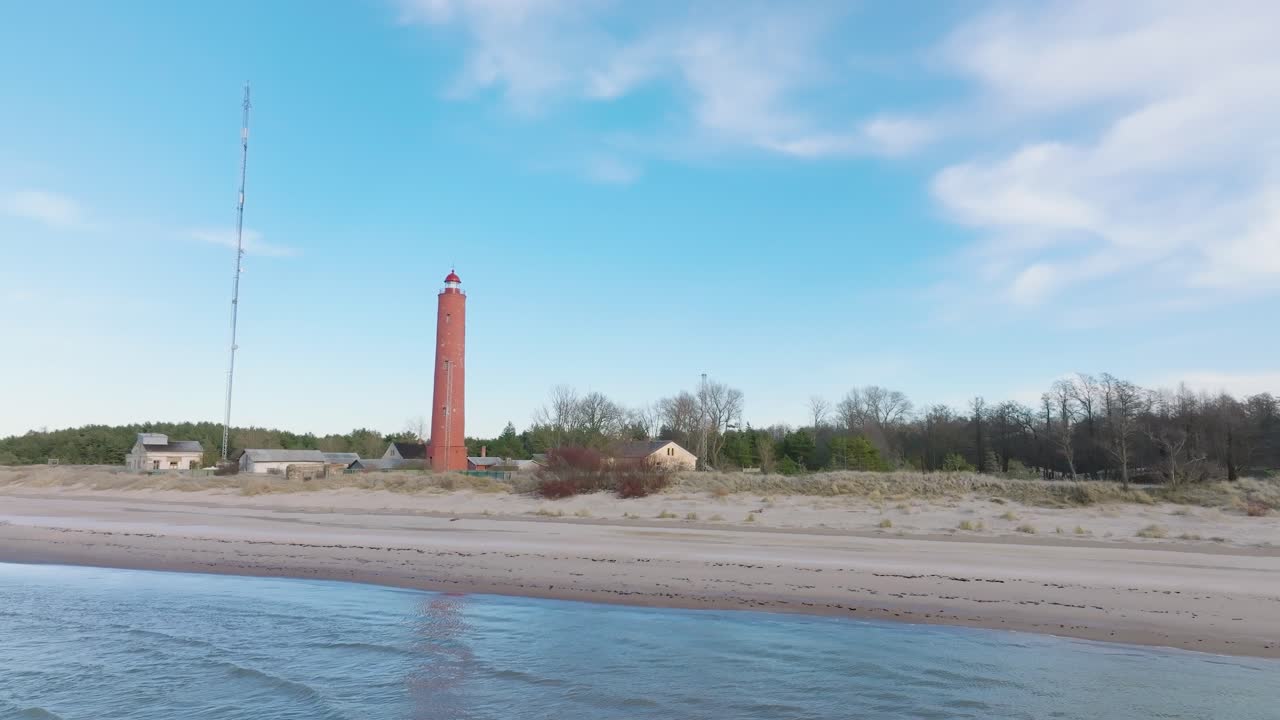 Aerial establishing view of red colored Akmenrags lighthouse, Baltic sea coastline, Latvia, white sand beach, calm sea, sunny day with clouds, distant wide drone shot moving forward low