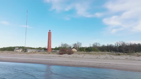 luftaufnahme des rot gefärbten leuchtturms von akmenrags, der ostseeküste, lettlands, weißem sandstrand, ruhiger see, sonniger tag mit wolken, weit entfernter drohnenschuss, der sich tief vorwärts bewegt