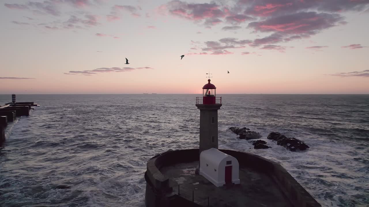 Red-and-white striped Farol da Barra lighthouse tower stands on rocky breakwater with beacon lit during golden hour dusk, Atlantic waters and horizon sky glowing amber