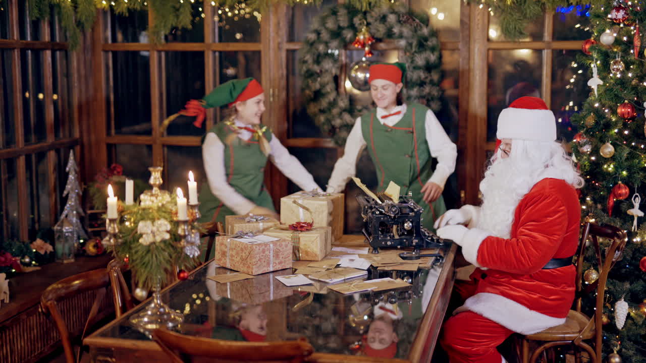 Little child is taking an envelope from Santa's residence on xmas background. Happy elves in fairy costumes dancing near Santa Claus who is typing at the table. Christmas atmosphere inside.