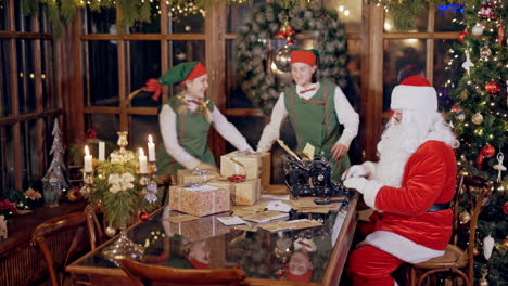 little child is taking an envelope from santa's residence on xmas background. happy elves in fairy costumes dancing near santa claus who is typing at the table. christmas atmosphere inside.