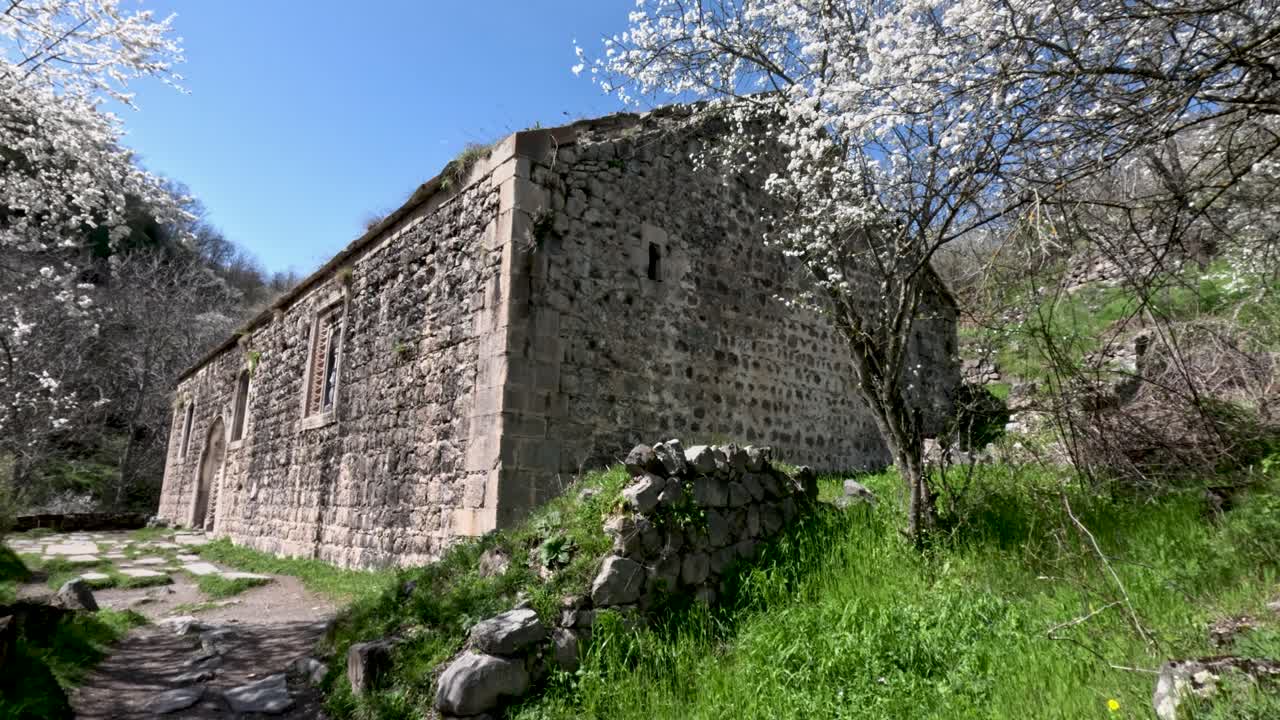 Una struttura in pietra si erge in un'area verde con alberi in fiore. Il sole splende luminoso in una giornata limpida, illuminando la natura circostante. Le persone possono esplorare il sito e godersi il panorama