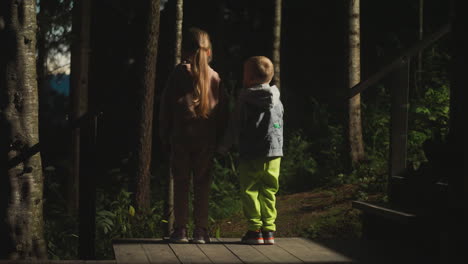 children stand on terrace wooden ground in dark forest. little brother and sister enjoy night woods beauty. kids observe park standing near cottage
