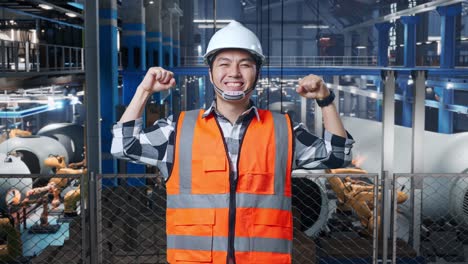 ingeniero asiático con casco de seguridad de pie en una fábrica de fabricación de turbinas eólicas, flexionando su bíceps y sonriendo a la cámara mientras el brazo robótico trabaja