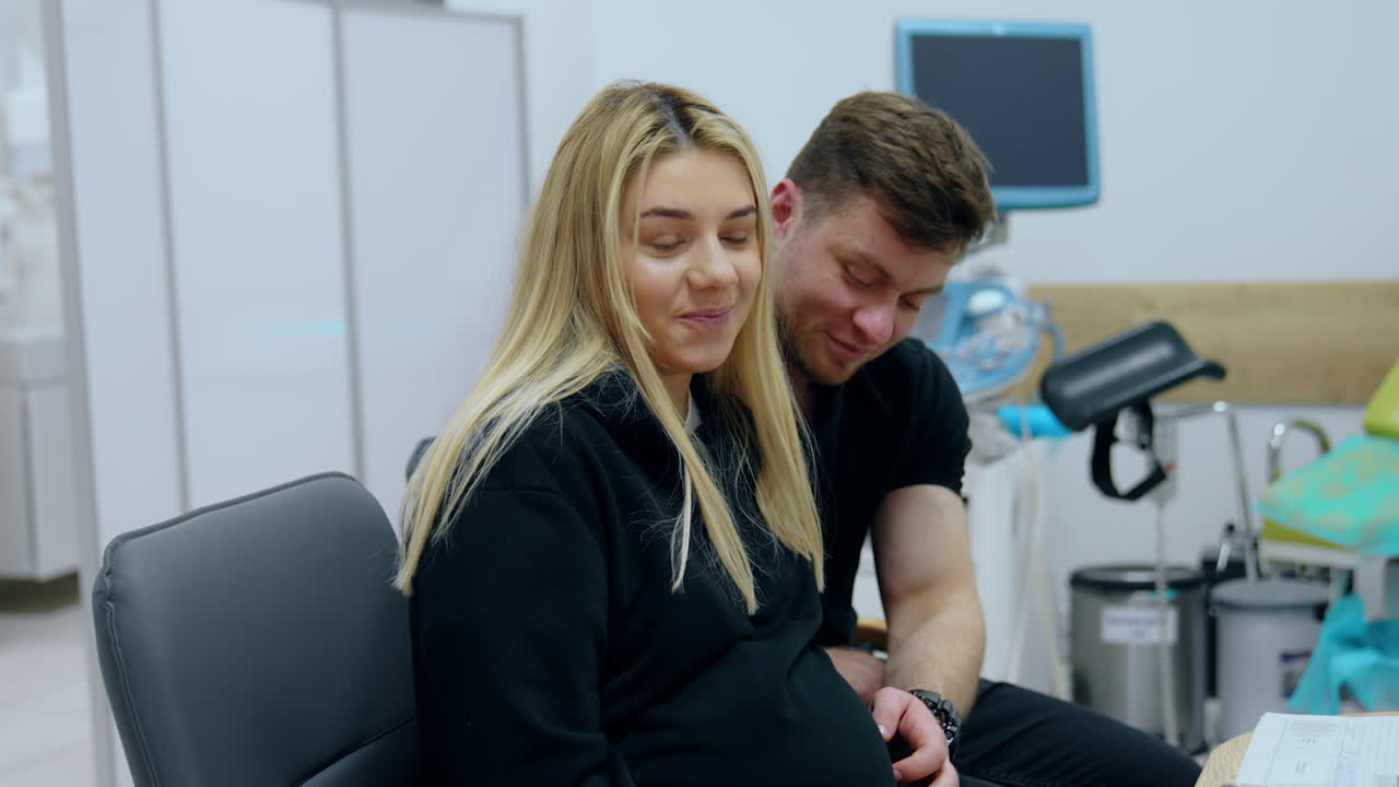 Caucasian couple expecting a baby is at the doctor's check up. Man strokes big wife's belly. Woman stands up to go to a ultrasound machine.