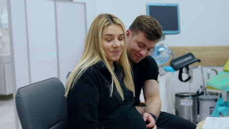 caucasian couple expecting a baby is at the doctor's check up. man strokes big wife's belly. woman stands up to go to a ultrasound machine.